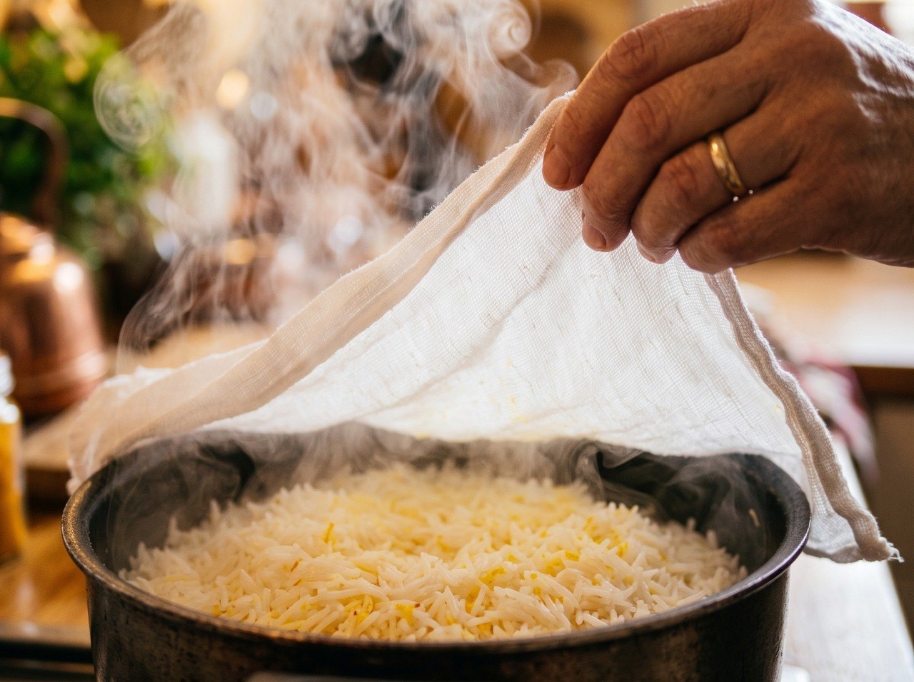 Person lifting a clean dry cloth from a pot of Persian rice to absorb excess steam, warm kitchen light, close up food photography